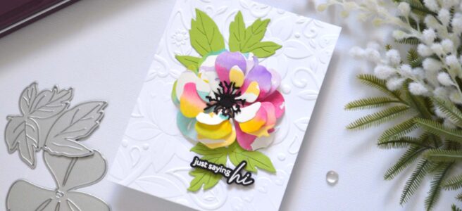 Handmade greeting card featuring a watercolor rainbow flower die cut layered with leaves, centered on a 3D embossed background, with the sentiment attached to the focal point.
