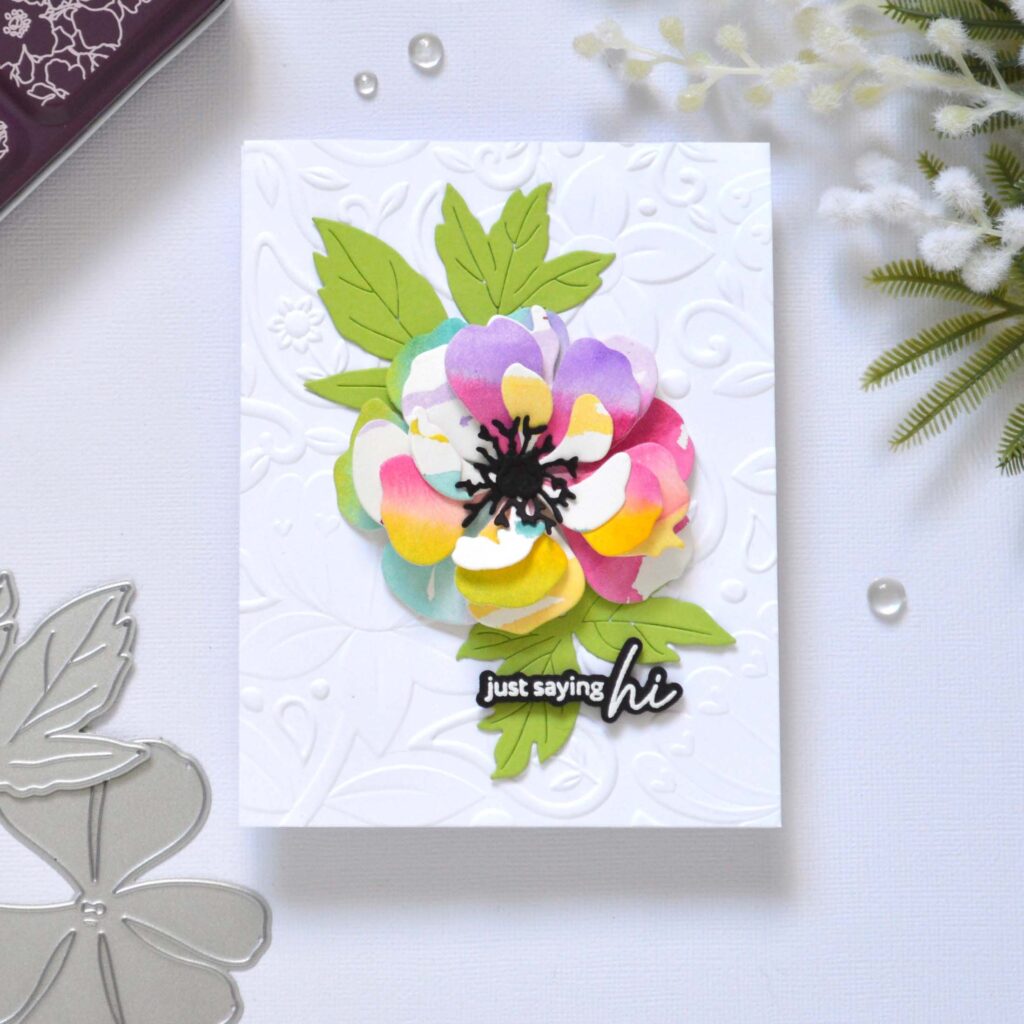 Handmade greeting card featuring a watercolor rainbow flower die cut layered with leaves, centered on a 3D embossed background, with the sentiment attached to the focal point.