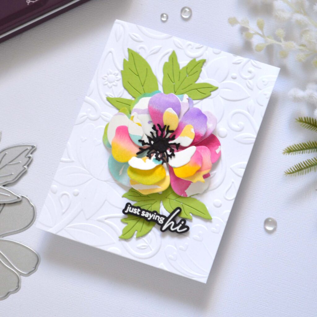 Handmade greeting card featuring a watercolor rainbow flower die cut layered with leaves, centered on a 3D embossed background, with the sentiment attached to the focal point.