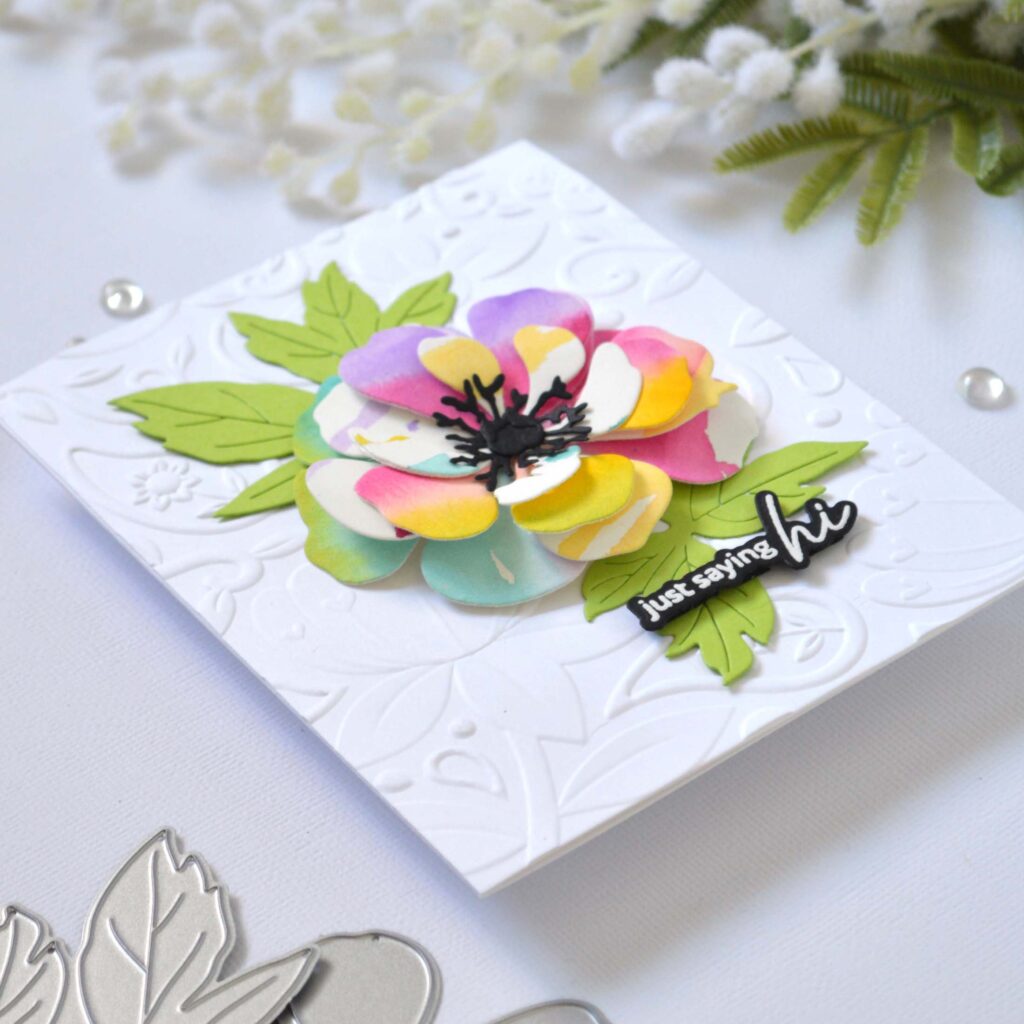 Handmade greeting card featuring a watercolor rainbow flower die cut layered with leaves, centered on a 3D embossed background, with the sentiment attached to the focal point.