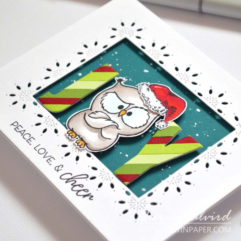 Humorous owl with Santa hat used as the “O” in JOY; letters die-cut from striped paper, owl in a decorative frame window over a teal snowy background.