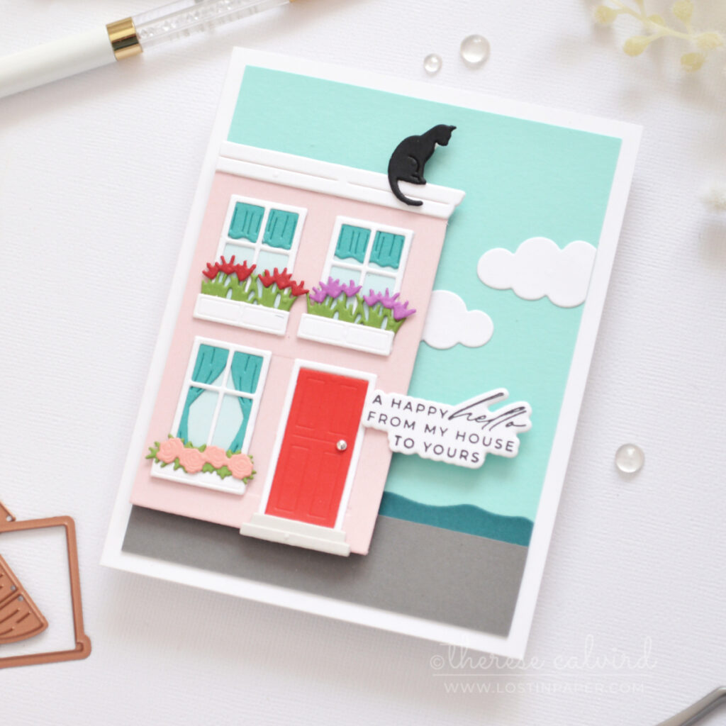 A2 handmade card featuring a trimmed Everyday City Holiday die cut pink house with flower boxes, rooftop cat, seaside scene and clouds by Therese Calvird