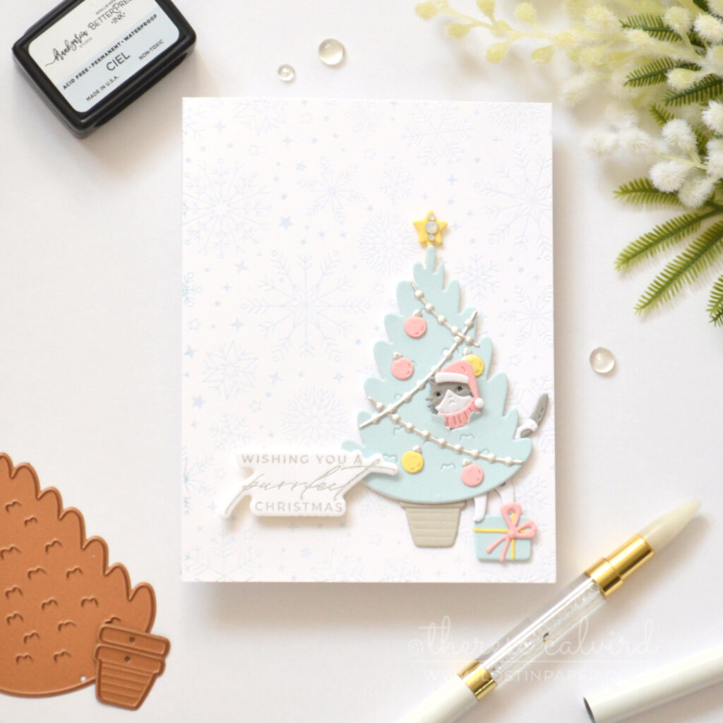 Grey cat peeking through a blue Christmas tree above a present on a pale blue snowflake background.
