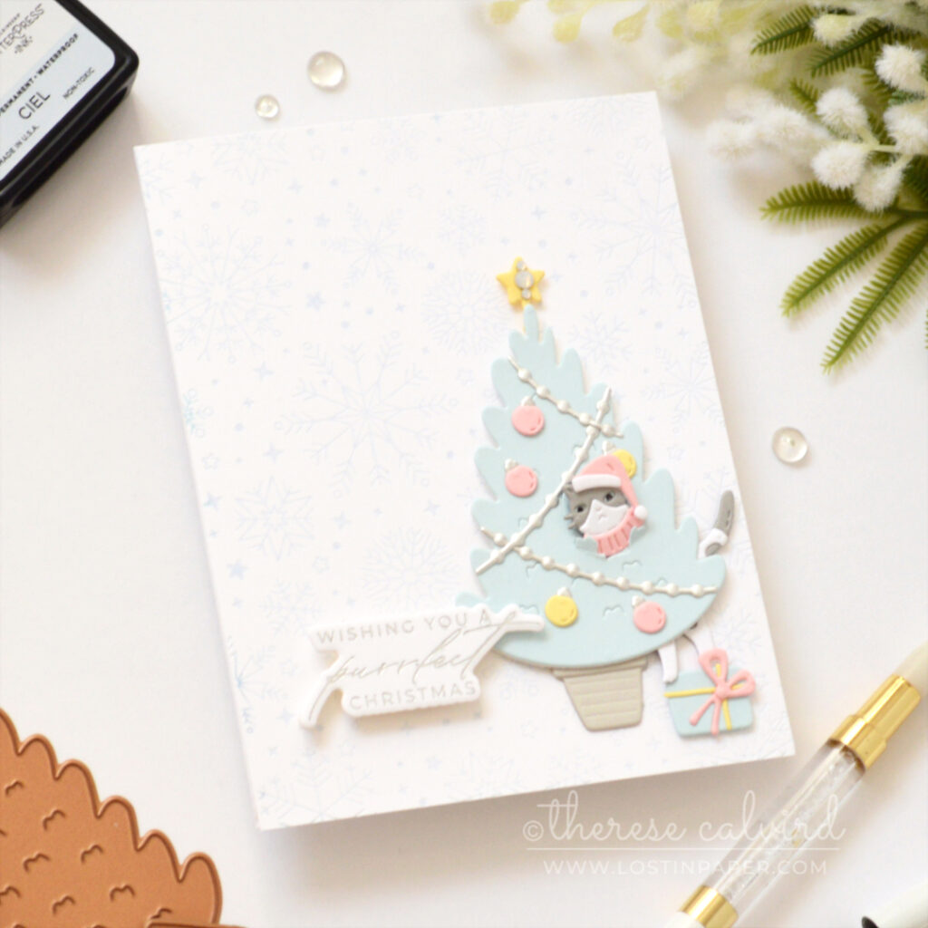 Grey cat peeking through a blue Christmas tree above a present on a pale blue snowflake background.
