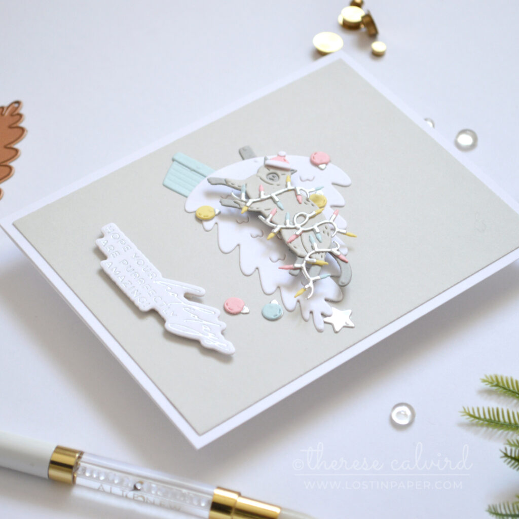 Grey cat tangled in Christmas lights while lying on a fallen white tree against a grey background.