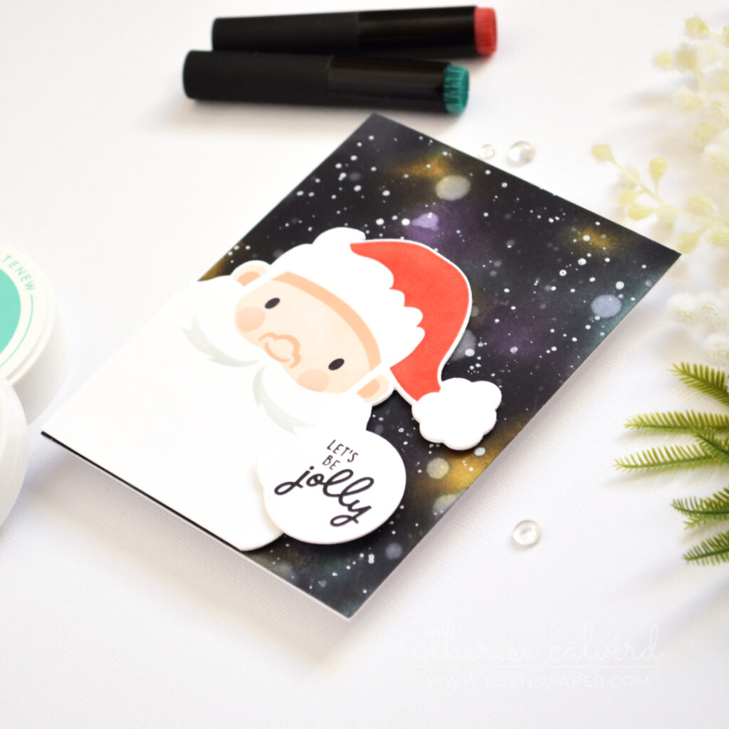 Christmas card with an ink-blended galaxy background, white splattered stars, and a large stenciled Santa face with a Jolly sentiment.