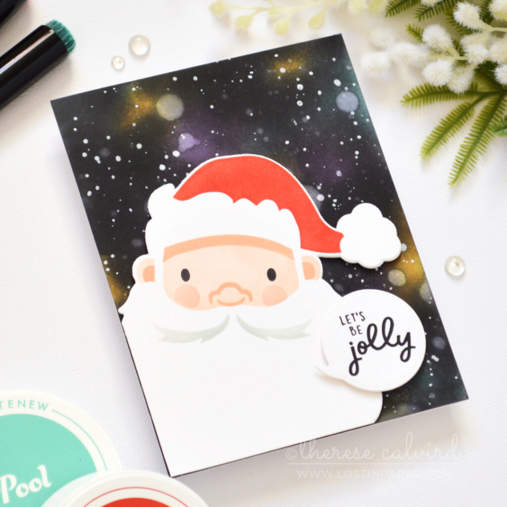 Christmas card with an ink-blended galaxy background, white splattered stars, and a large stenciled Santa face with a Jolly sentiment.