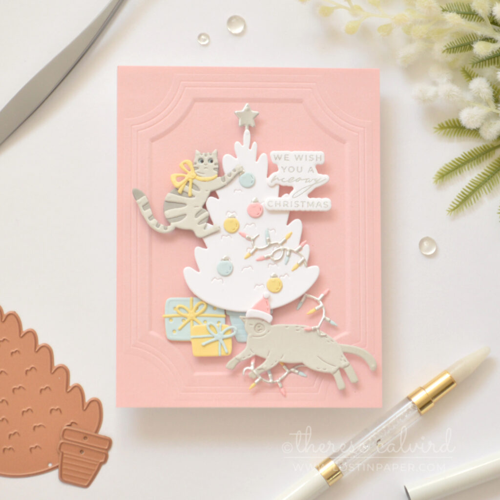 Grey die-cut cat climbing a white Christmas tree on a pink background with pastel holiday decorations.