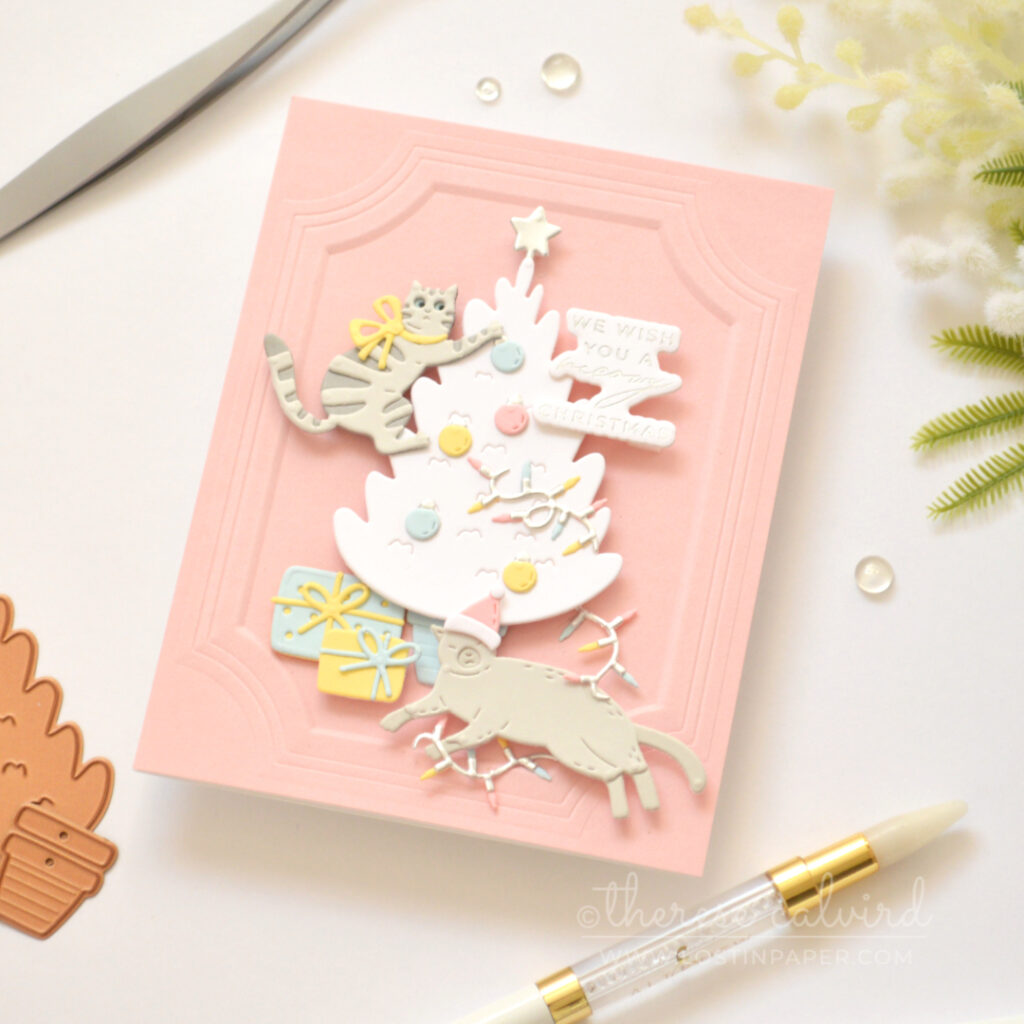 Grey die-cut cat climbing a white Christmas tree on a pink background with pastel holiday decorations.