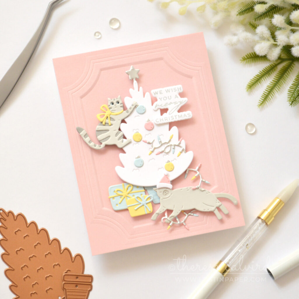 Grey die-cut cat climbing a white Christmas tree on a pink background with pastel holiday decorations.