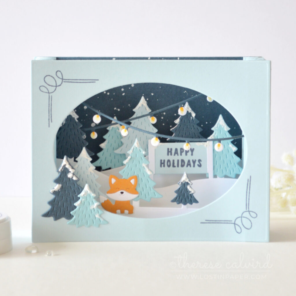 Blue accordion Christmas card with layered snowy trees, holiday lights and an orange fox by Therese Calvird