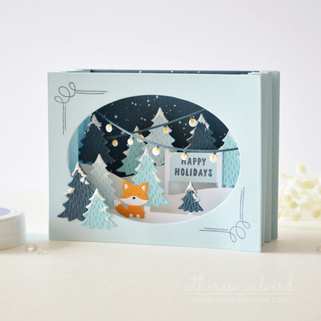 Blue accordion Christmas card with layered snowy trees, holiday lights and an orange fox by Therese Calvird