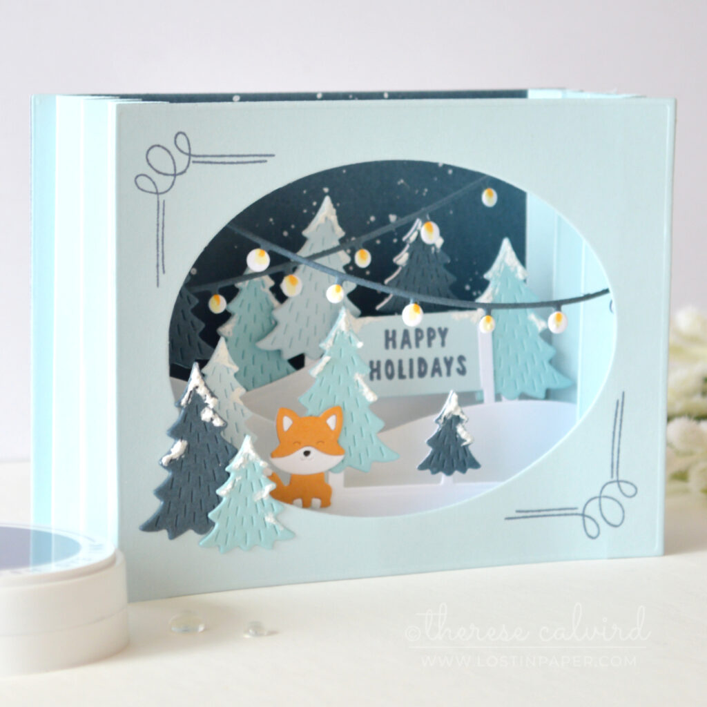 Blue accordion Christmas card with layered snowy trees, holiday lights and an orange fox by Therese Calvird