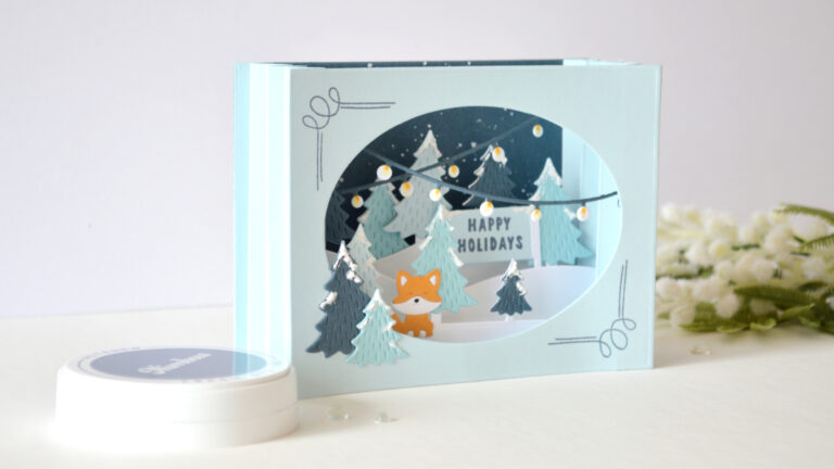 Blue accordion Christmas card with layered snowy trees, holiday lights and an orange fox by Therese Calvird