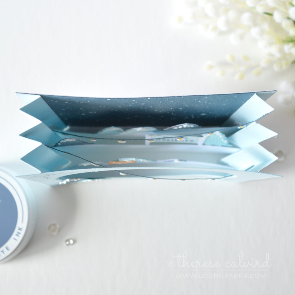 Blue accordion Christmas card with layered snowy trees, holiday lights and an orange fox by Therese Calvird