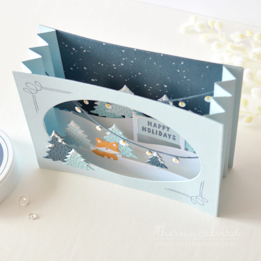 Blue accordion Christmas card with layered snowy trees, holiday lights and an orange fox by Therese Calvird