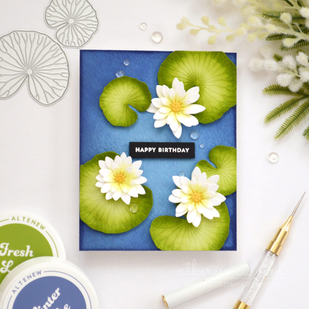 Handmade pond lilies birthday card with white and lime flowers, green lily pads, gold centres and water droplets on a textured blue background.