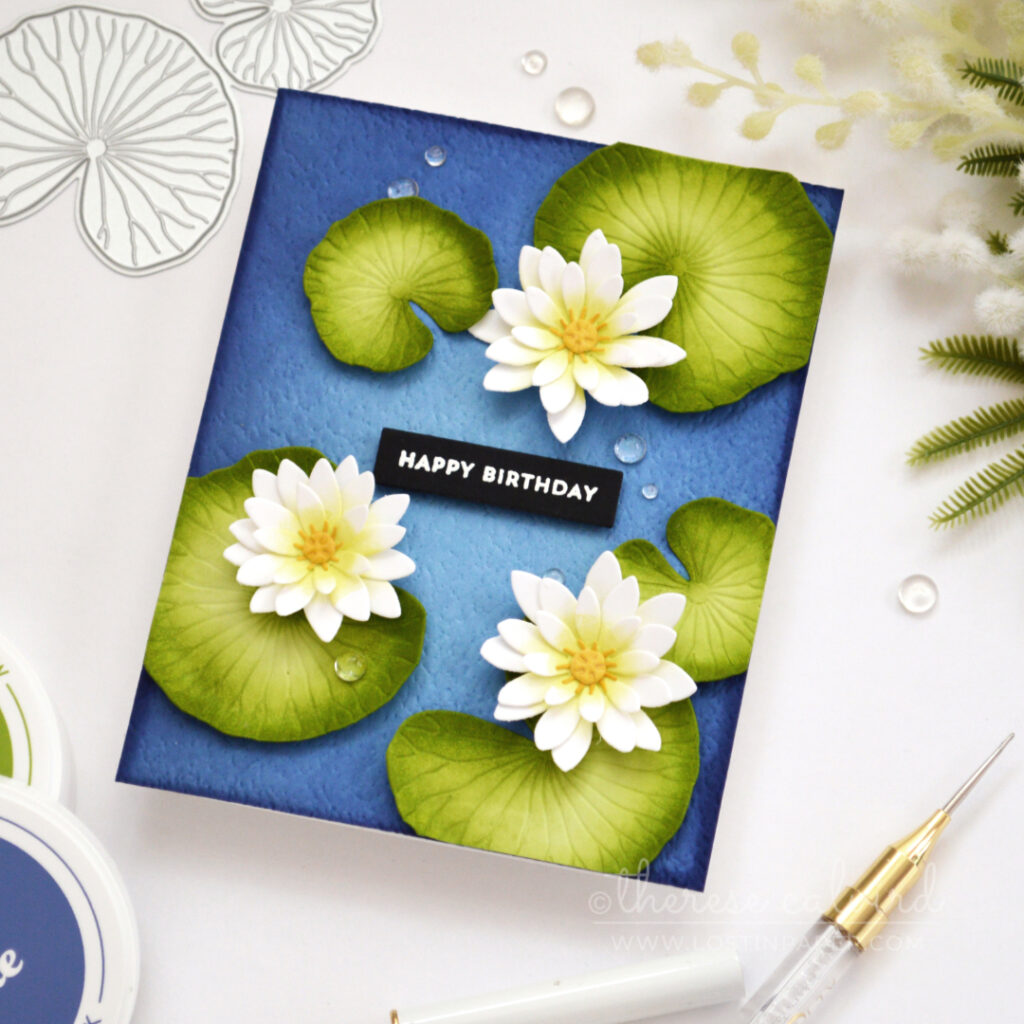Handmade pond lilies birthday card with white and lime flowers, green lily pads, gold centres and water droplets on a textured blue background.