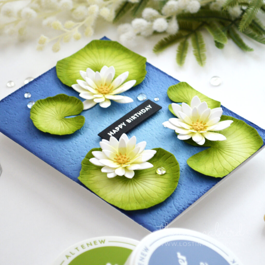 Handmade pond lilies birthday card with white and lime flowers, green lily pads, gold centres and water droplets on a textured blue background.