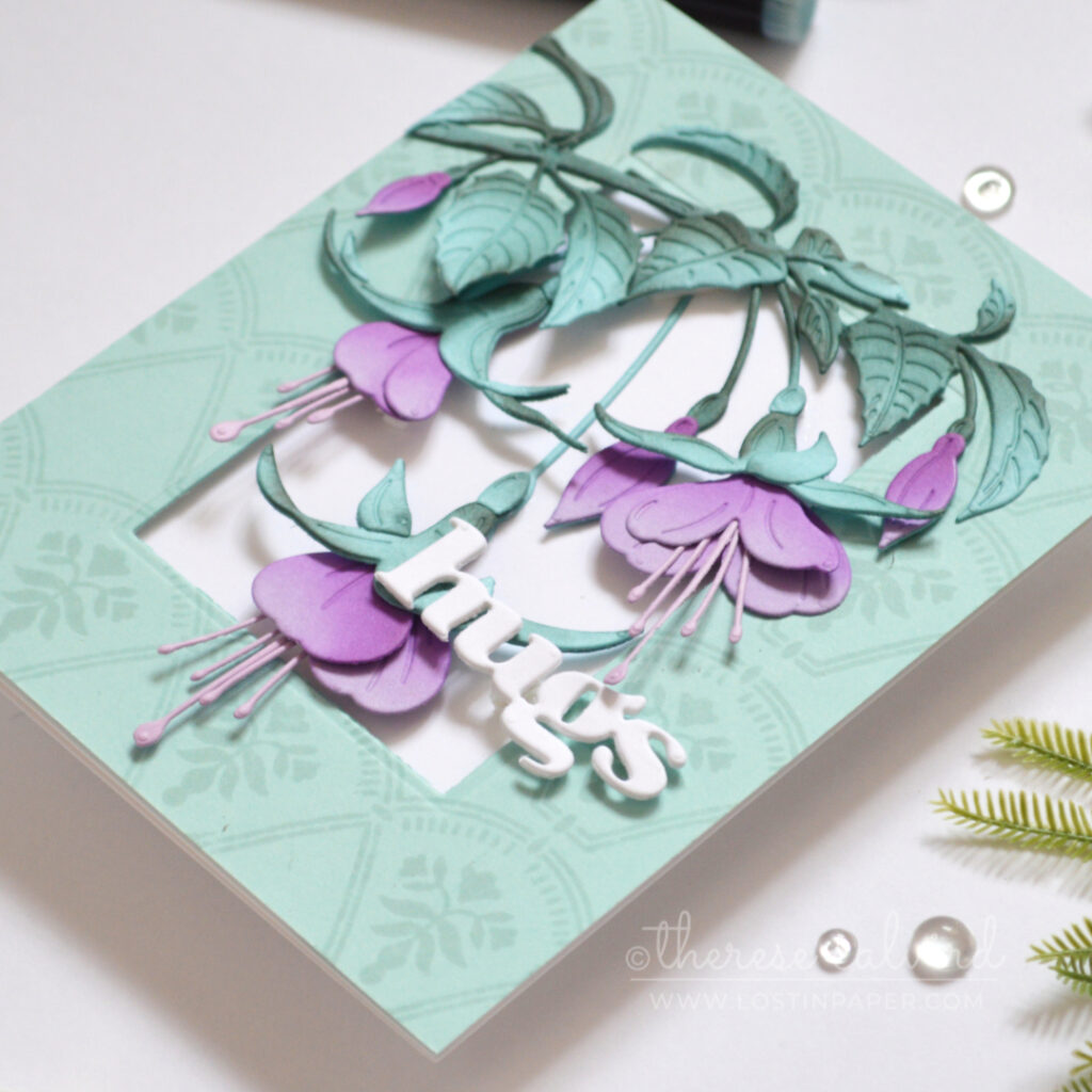 Handmade card with purple fuchsia flowers and teal leaves on a pale teal tone-on-tone stamped frame, finished with a white die-cut “hugs” sentiment.