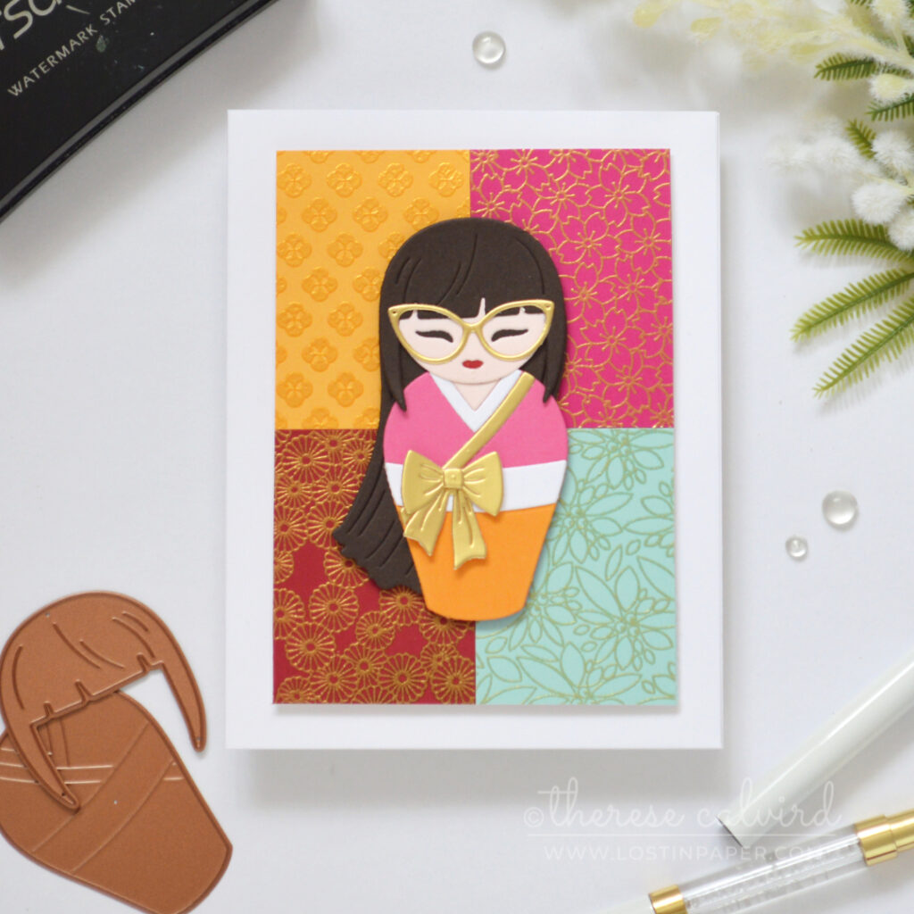 Die cut Kokeshi girl on a colourful grid of gold embossed kimono patterns.