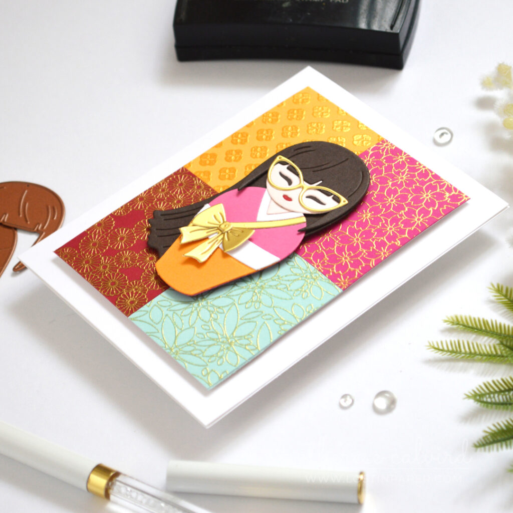 Die cut Kokeshi girl on a colourful grid of gold embossed kimono patterns.