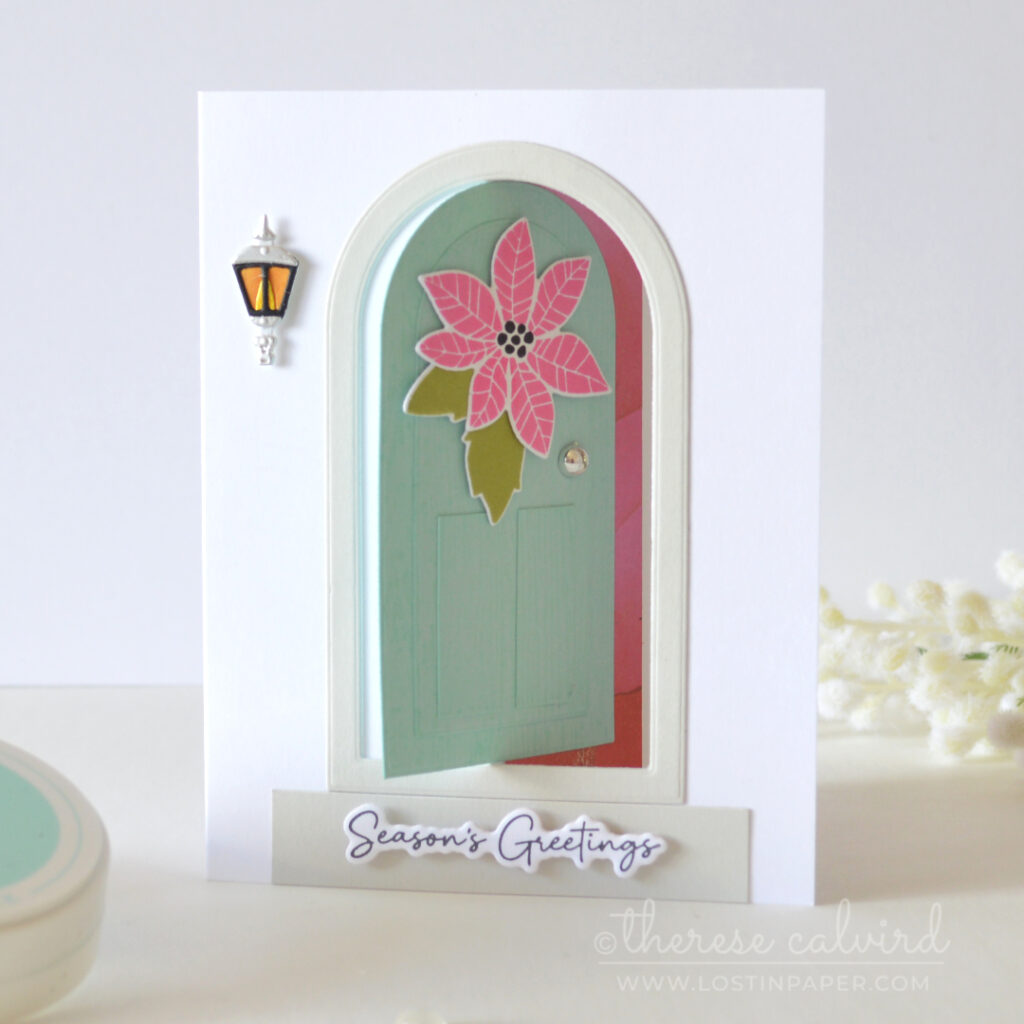 Interactive fun fold Christmas card with a light teal door, pink poinsettia, green leaves, lantern and silver door knob.