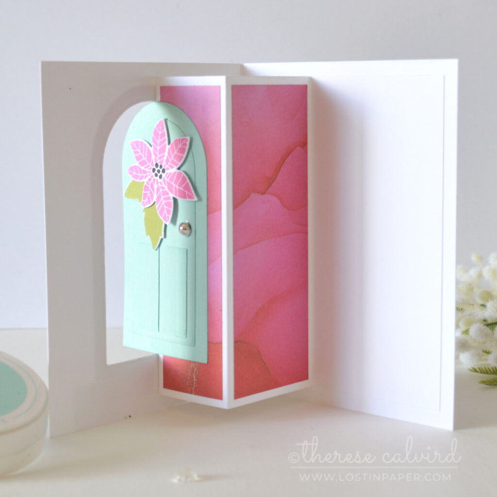 Interactive fun fold Christmas card with a light teal door, pink poinsettia, green leaves, lantern and silver door knob.