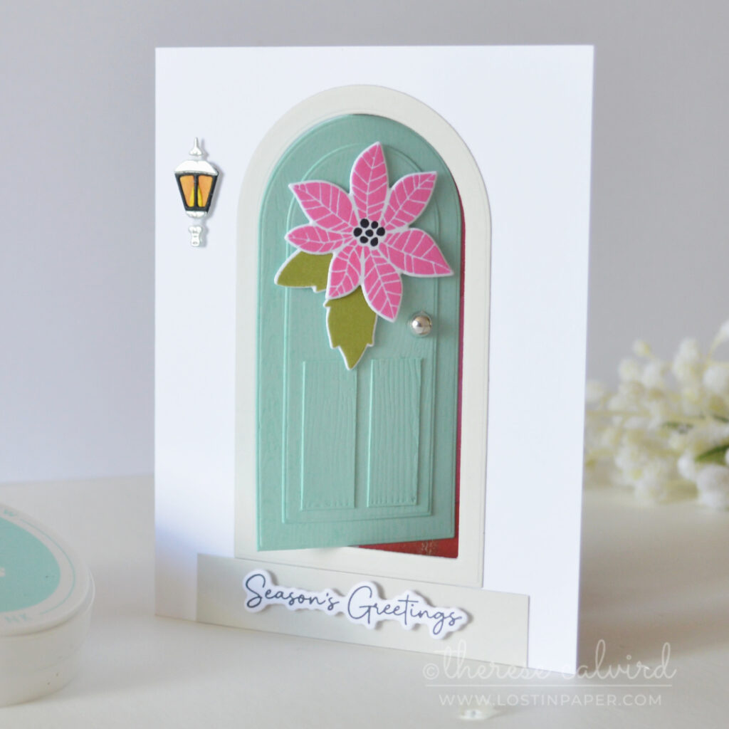 Interactive fun fold Christmas card with a light teal door, pink poinsettia, green leaves, lantern and silver door knob.